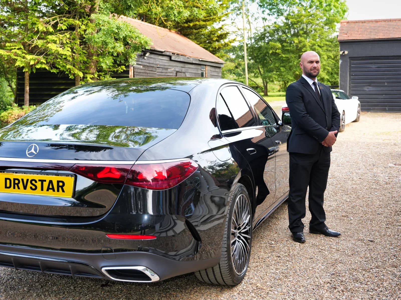 A DRVSTAR chauffeur in a dark suit standing beside a black Mercedes-Benz E-Class