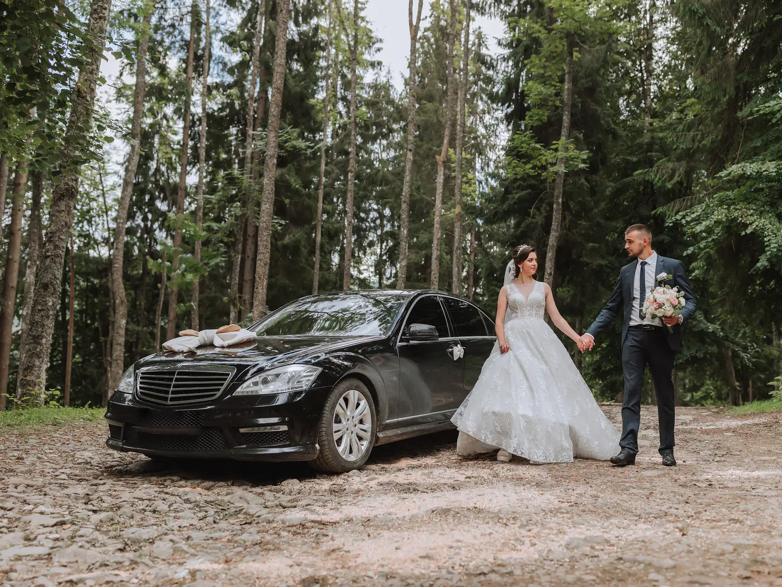 A bride and groom beside a black Mercedes with a wedding ribbon on a forest road