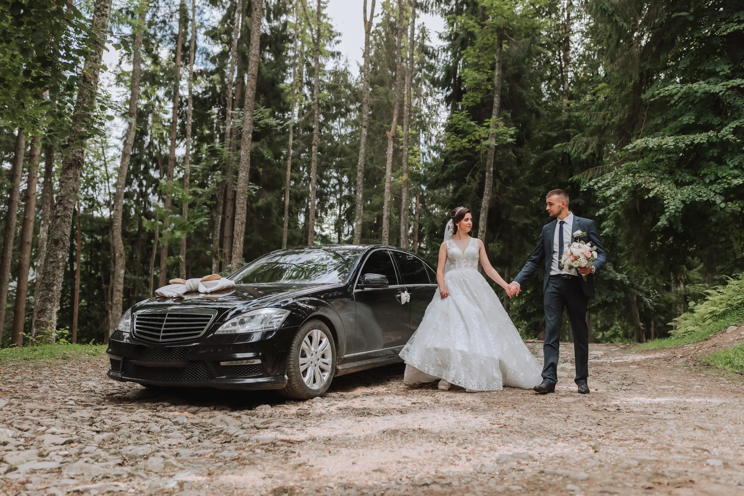 Bride and groom holding hands beside a black Mercedes wedding car on a tree-lined drive