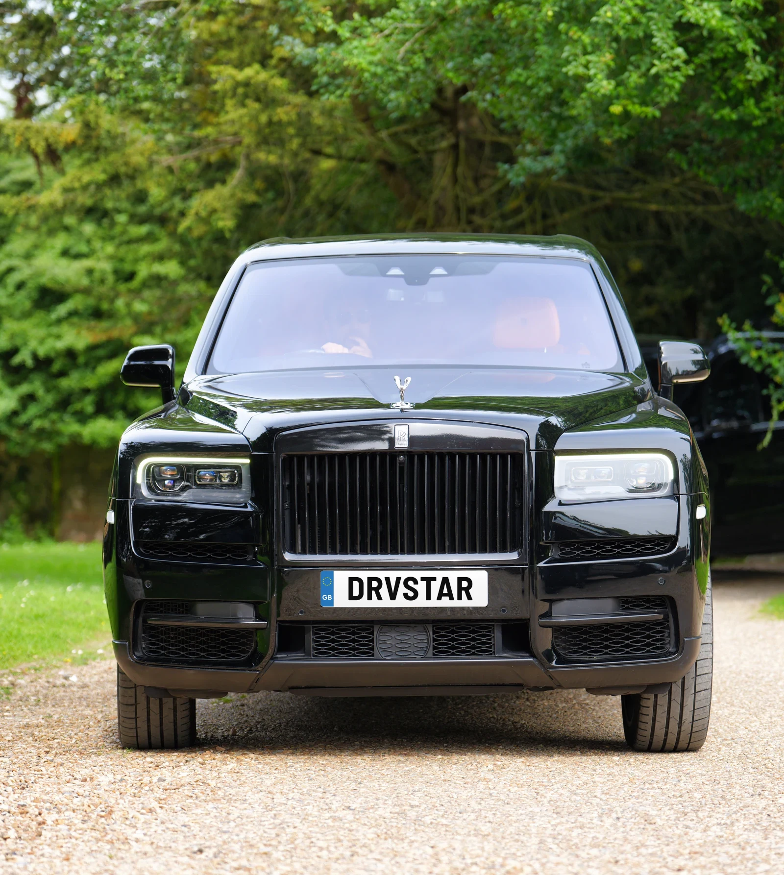 A black Rolls-Royce Cullinan with a UK number plate on a gravel country drive, framed by trees and a lamp post
