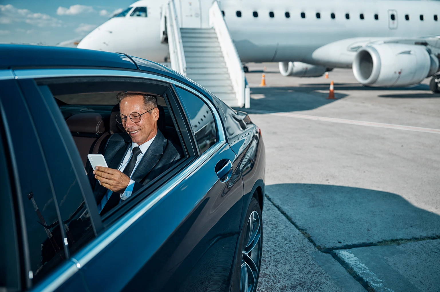 Business traveller checking his phone in a chauffeur-driven car beside a private jet on the tarmac