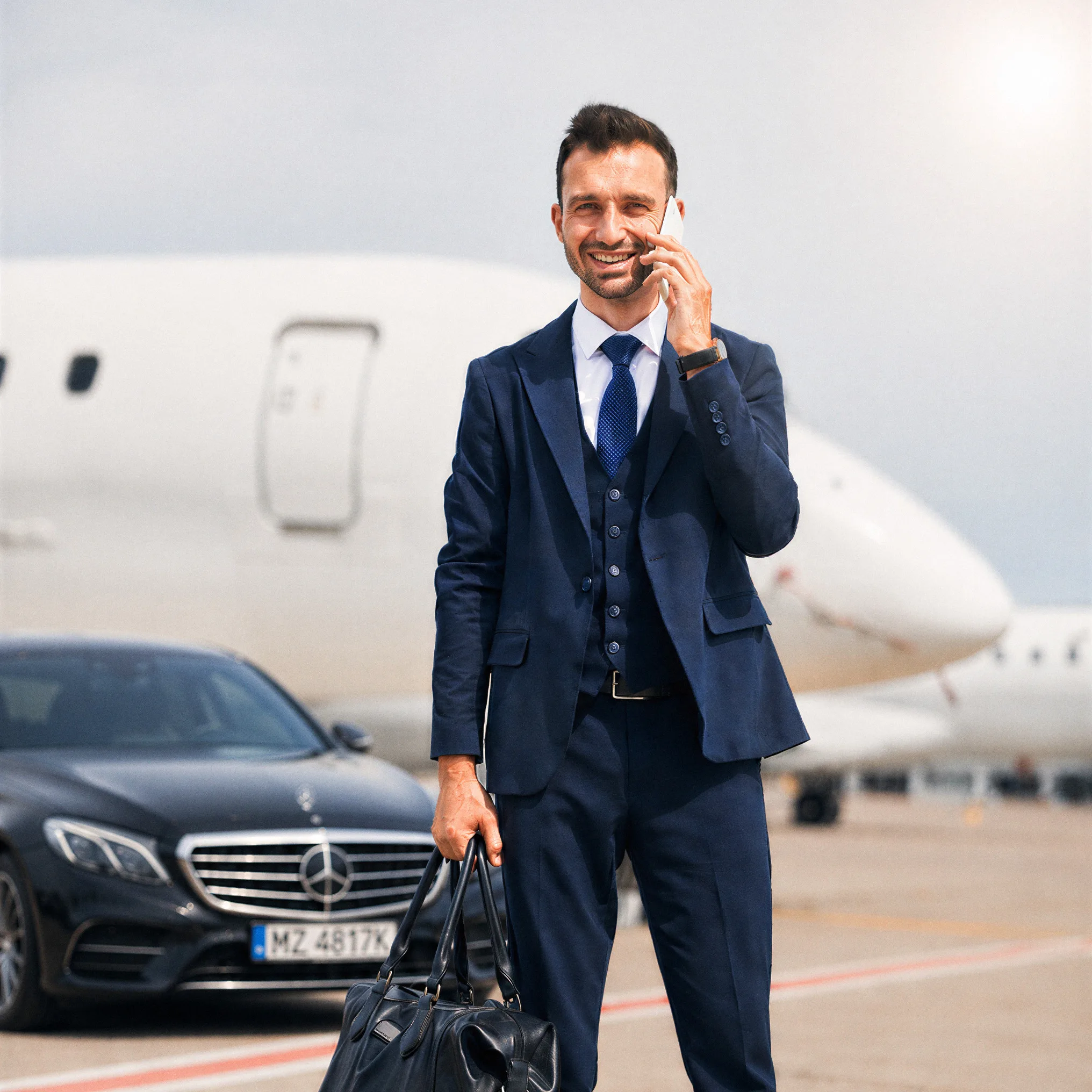 Traveller on a phone call with his holdall beside a chauffeur-driven Mercedes and private jet on the tarmac