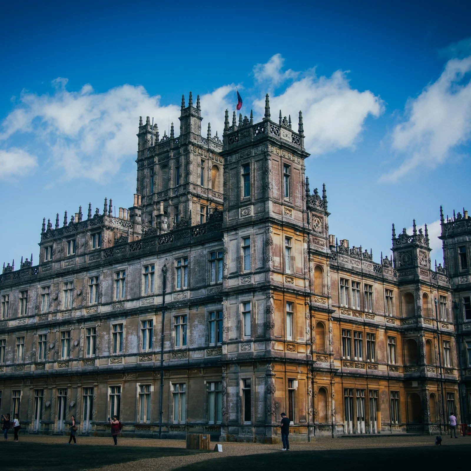 Highclere Castle in afternoon sun, its Victorian towers and pinnacles against a blue sky with visitors on the lawn